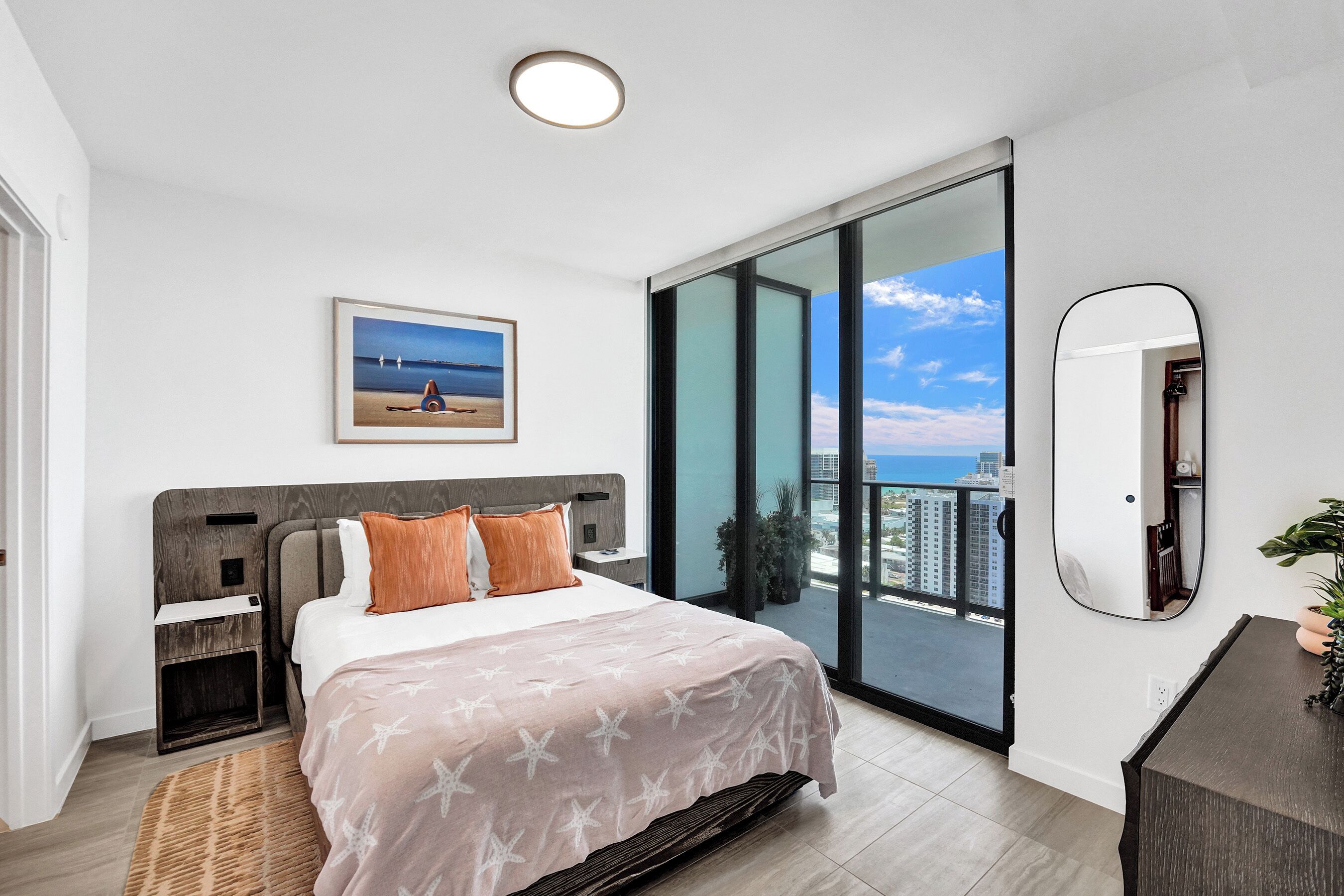Un dormitorio con una cama grande, una mesita de noche, un aparador, un espejo y vistas a la ciudad a través de ventanales del suelo al techo.