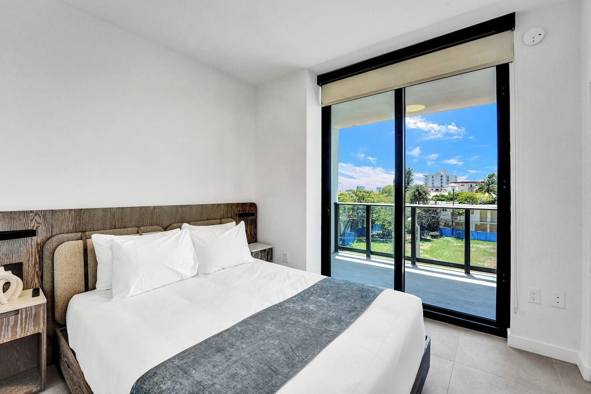 Ein modernes Hotelzimmer mit einem großen Bett, einem hölzernen Kopfteil, einem Nachttisch und einem Balkon mit Blick auf die Stadt.