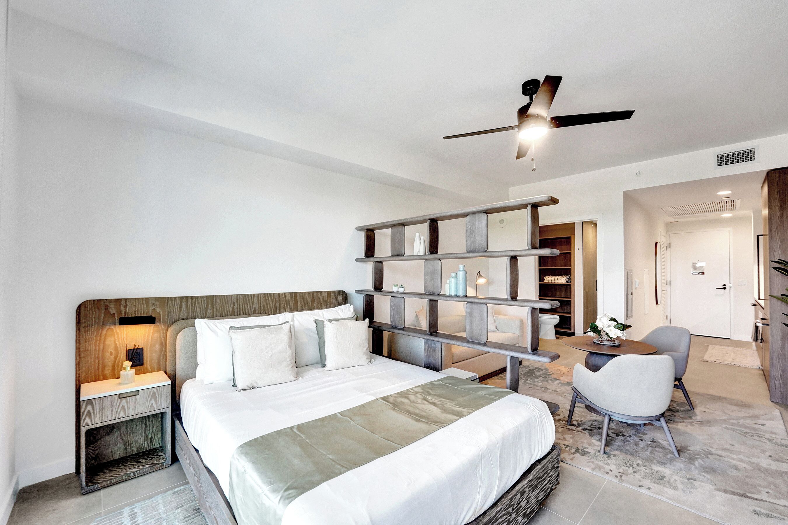 Une chambre d’hôtel moderne dotée d’un grand lit, d’une table de chevet, d’un coin salon avec une table et des chaises, et d’une bibliothèque intégrée en bois.