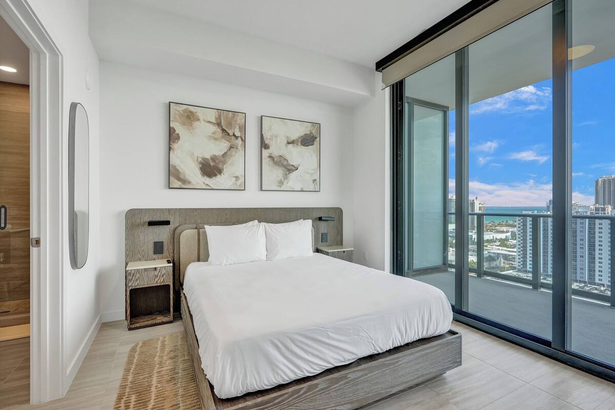 Ein modernes Schlafzimmer mit einem großen Bett, zwei an Wänden befestigten abstrakten Gemälden, einem Nachttisch und Blick auf eine Skyline durch raumhohe Fenster.