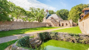 A courtyard with a stone wall, a grassy area, a small pond, and a building with a staircase and a tower.