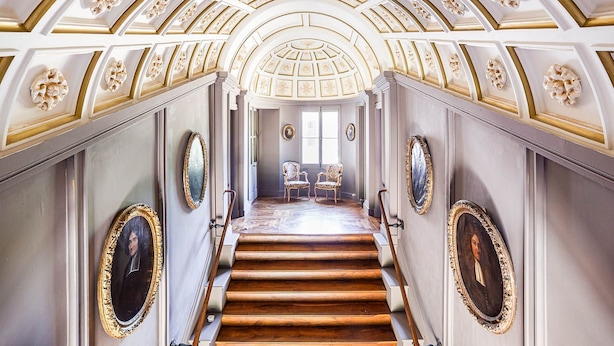 A grand staircase with ornate gold detailing, leading to a room with two chairs and a large window.