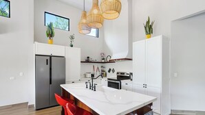 A modern kitchen with white cabinetry, a stainless steel refrigerator, a central island with a sink, and red bar stools. - Villa with Minimalist Design & Scenic Deck with Private Infinity Pool and Jungle Views (Coco)
