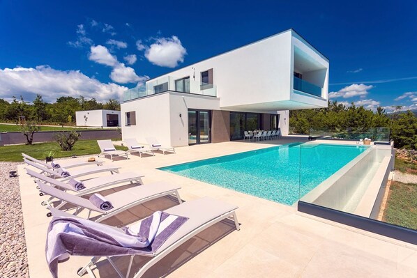 Une maison moderne dotée d’une piscine, de chaises longues et d’une terrasse fermée par des baies vitrées.