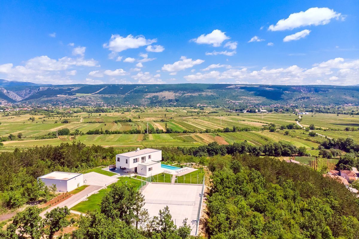 A modern two-story house with a swimming pool, surrounded by lush green fields and mountains in the background.