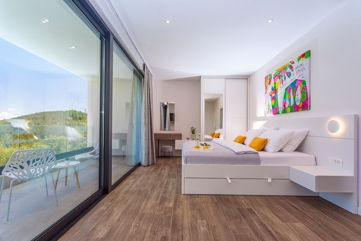 A modern bedroom with a large bed, a desk, and a balcony with glass doors.