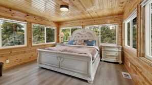 Dormitorio de una cabaña de madera con una cama grande, una mesita de noche y una ventana con vistas a un bosque.
