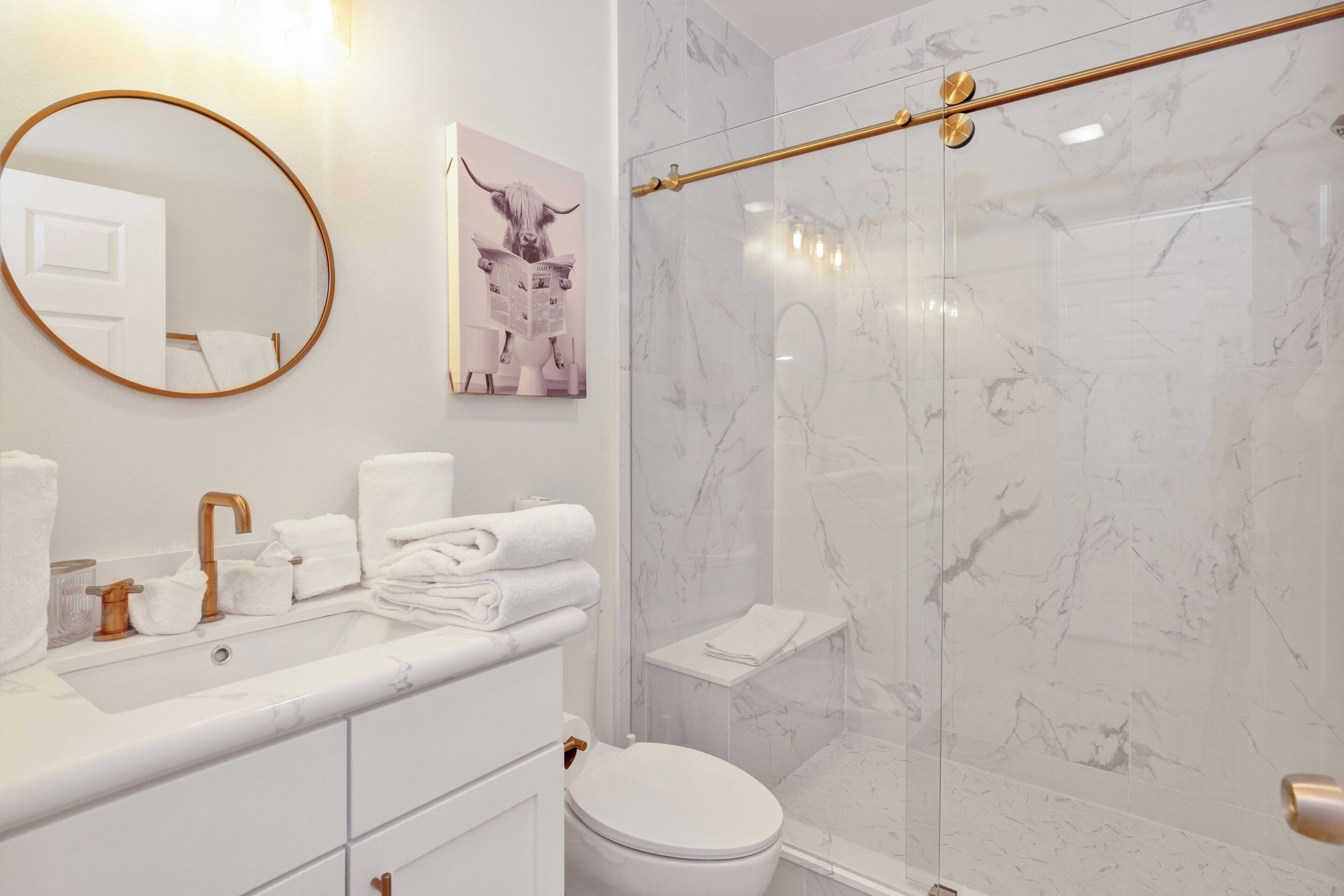Un baño moderno con ducha de mármol, espejo redondo y lavamanos blanco con detalles dorados.