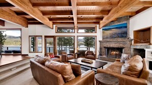 A spacious living room with a stone fireplace, leather sofas, and large windows overlooking a lake. - Bear Lake Lodge #2547 (Big Bear Lake)