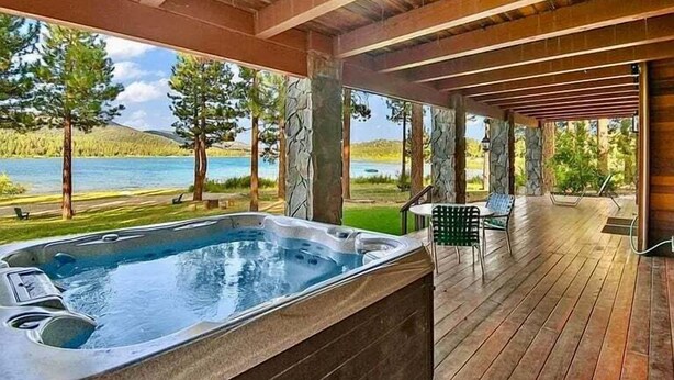 A hot tub area with a wooden deck, stone pillars, and a view of a lake and trees. - Bear Lake Lodge #2547 (Big Bear Lake)