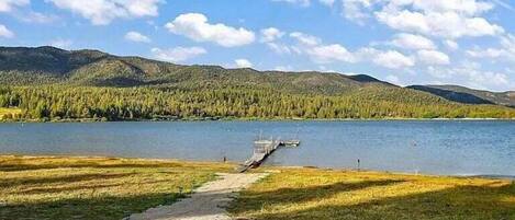 A lakeside view with a dock extending into the water, surrounded by grassy areas and forested hills.