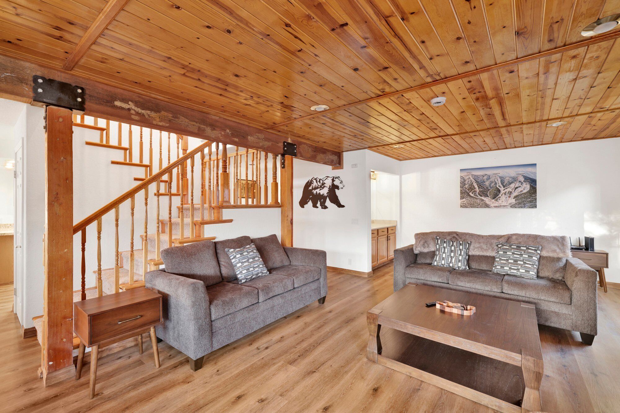 A spacious living area with wooden floors, two sofas with patterned cushions, a coffee table, a wooden staircase, and a wall-mounted bear decoration.