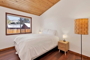 Un dormitorio con techo de madera, un ventanal con vistas a un paisaje nevado, una cama con sábanas blancas, una mesita de noche con una lámpara y una lámpara de pie de fibra trenzada.