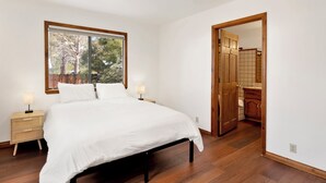 Un dormitorio con una cama grande, una mesita de noche, una lámpara, una ventana con vistas a árboles, y una puerta de madera que conduce a un baño.