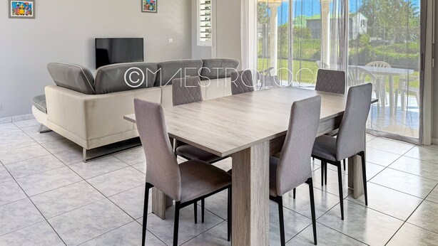 A modern living room with a sofa, dining table, and chairs. There is a TV mounted on the wall, and a window with a view of greenery outside. - Villa Perrin Évasion à 9 min de l'aéroport de la Guadeloupe (Les Abymes)