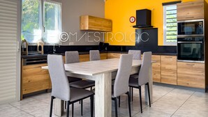 A modern kitchen with a dining area, featuring wooden cabinets, a black stove, and a large window with blinds. - Villa Perrin Évasion à 9 min de l'aéroport de la Guadeloupe (Les Abymes)