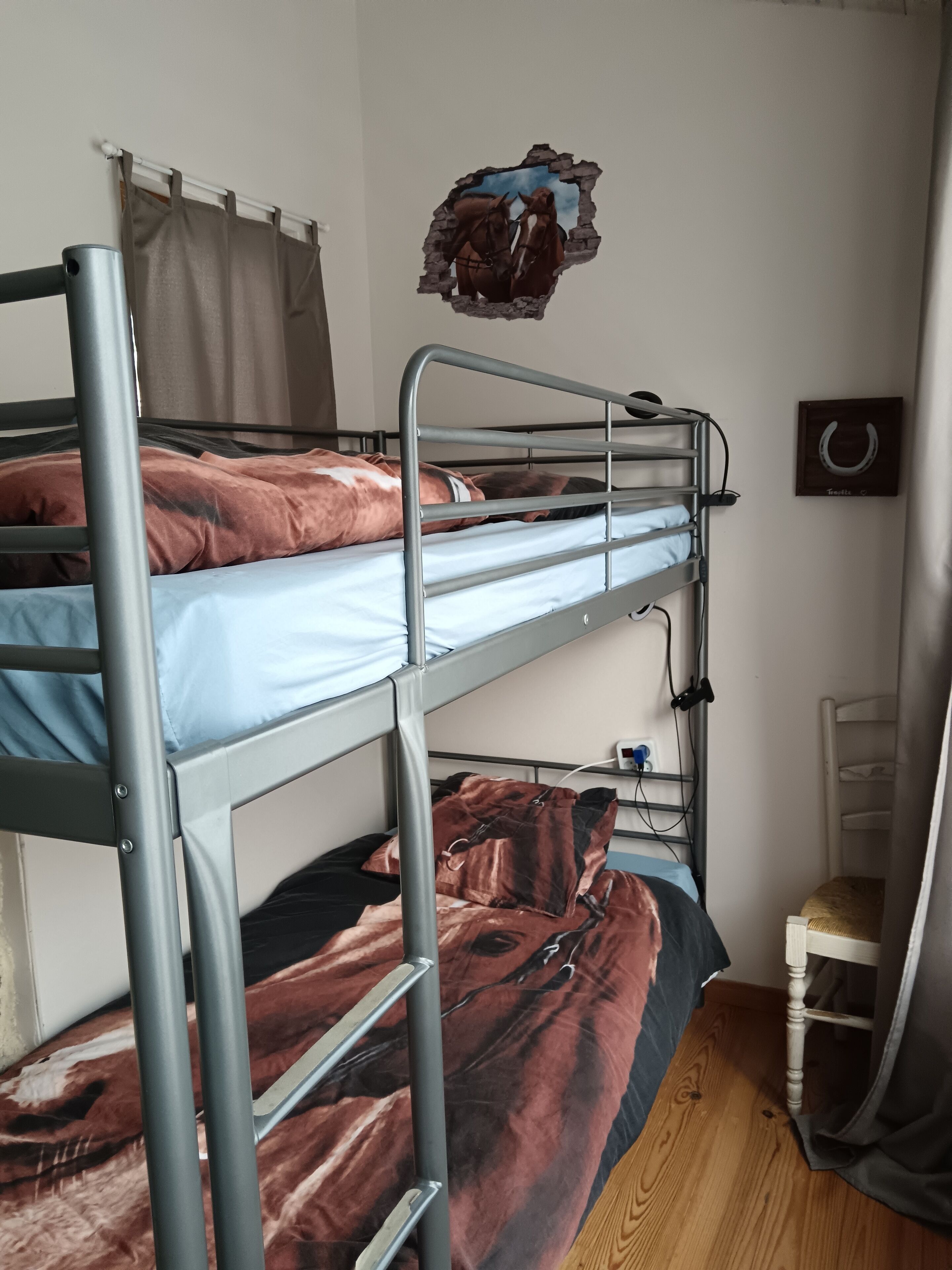 Une chambre avec un lit superposé, un rideau, un élément décoratif mural et une petite chaise en bois.