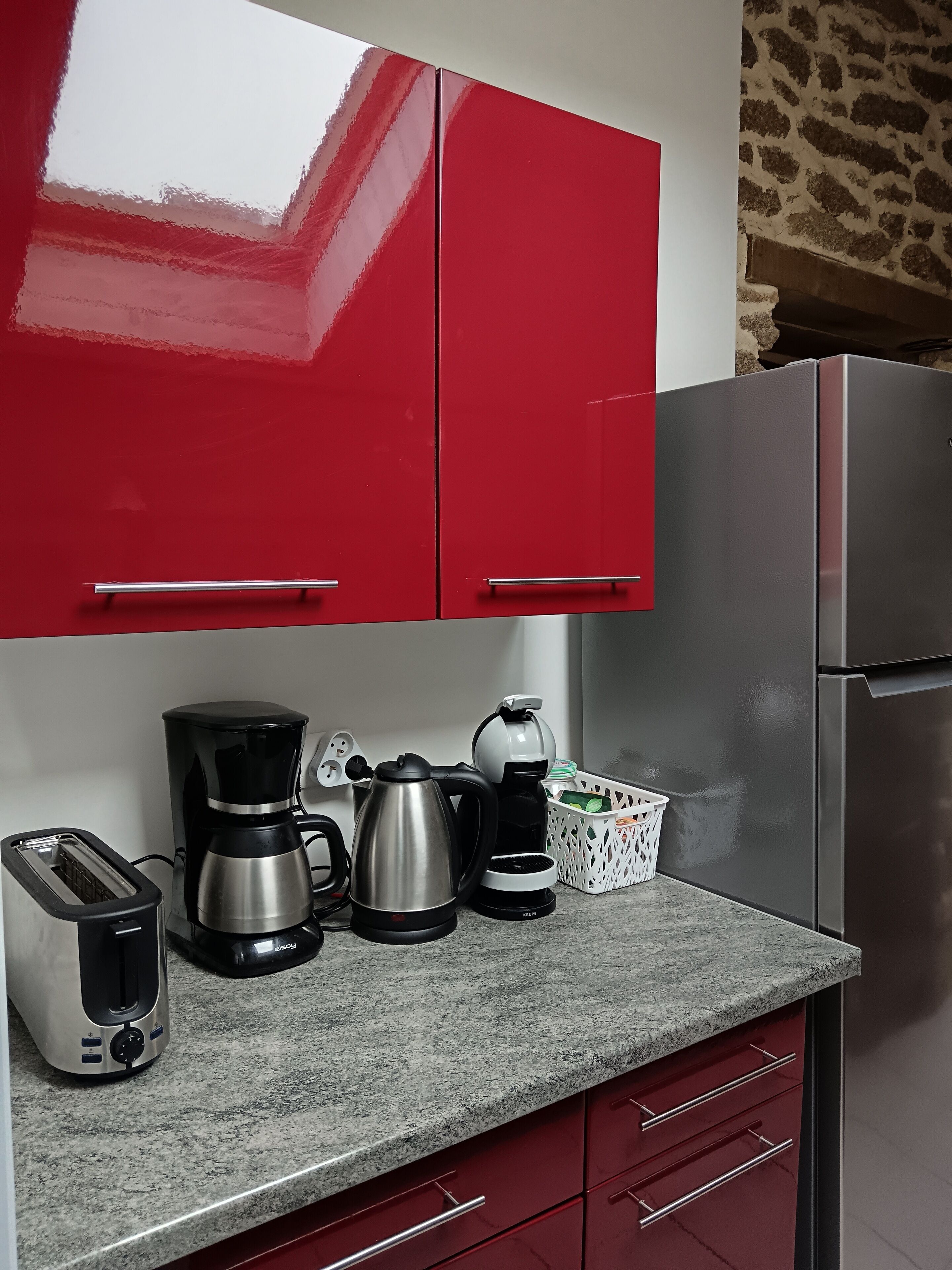 A kitchen with red cabinets, a stainless steel refrigerator, a coffee maker, a kettle, and a toaster.