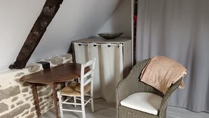 Un área de comedor con una mesa de madera y una silla blanca, una silla de mimbre con cojín, una pared de piedra y una mesita con un cuenco.