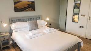 Una cama bien hecha con sábanas blancas y dos almohadas, una lámpara de noche y un cuadro con un paisaje en la pared.