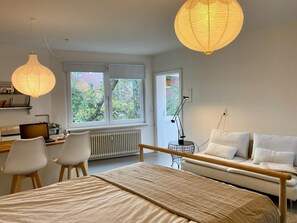 A modern bedroom with a bed, a desk with a computer, two chairs, a sofa, and a window with a view of trees.