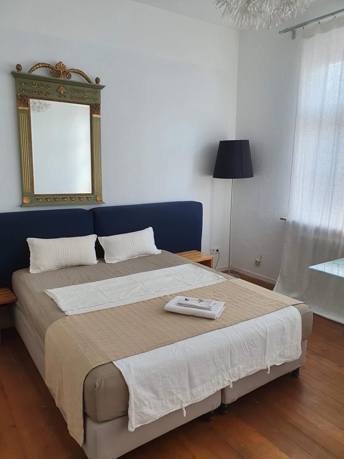 Una cama bien hecha con colcha color beige y blanco, un espejo grande con detalles ornamentales dorados, un cabecero azul oscuro y una lámpara de noche.