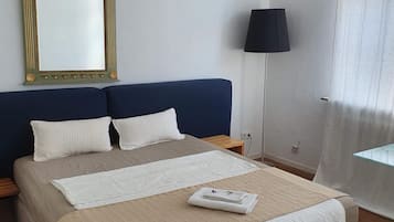 Una cama bien hecha con colcha color beige y blanco, un espejo grande con detalles ornamentales dorados, un cabecero azul oscuro y una lámpara de noche.
