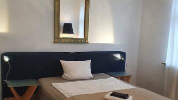 Uma cama bem arrumada com cabeceira, mesa de cabeceira com abajur, uma cadeira e um espelho grande na parede.
