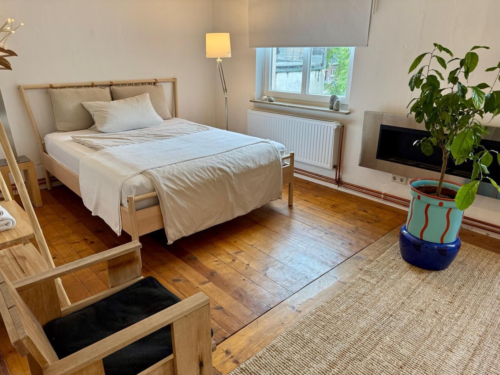 A bedroom with a bed, bedside table, lamp, radiator, window, TV, and a potted plant.