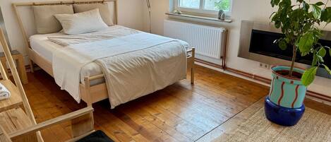 A bedroom with a bed, bedside table, lamp, radiator, window, TV, and a potted plant.