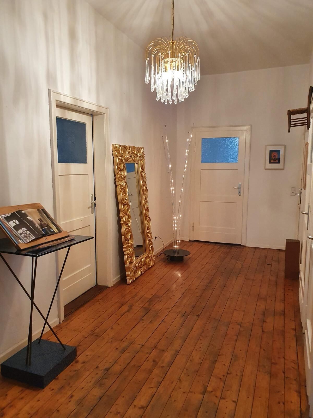 Une pièce avec un plancher en bois, deux portes blanches, un lustre et une table sur laquelle est posée une photo encadrée.