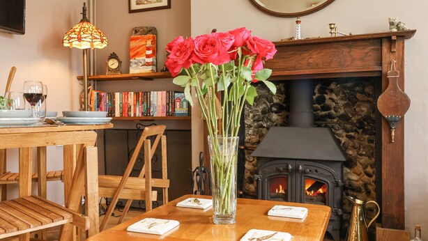 A cozy living room with a fireplace, a wooden table with a vase of flowers, a bookshelf, and a mirror. - 1 Rumsby Cottages (Lowestoft)