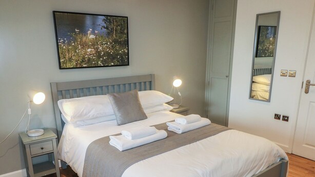 A neatly made bed with white linens and a grey headboard, a bedside table with a lamp, and a framed picture of a natural scene on the wall. - Chestnut (Cowbridge)