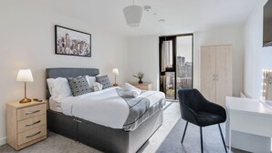 A modern hotel room with a bed, bedside tables, a desk, a chair, a wardrobe, and a city view. - Skyline Views | 2 Bed 2 Bath in Prime Location | Parking/Netflix! (Birmingham)