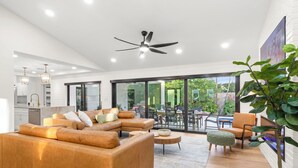 A modern living room with a sectional sofa, a dining area with outdoor seating, and a kitchen in the background.