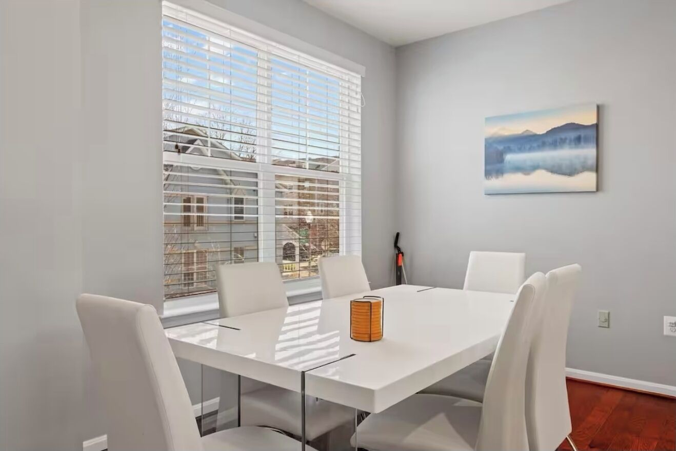 Un espace repas moderne avec une table et des chaises blanches, de grandes fenêtres équipées de stores et un tableau accroché au mur.
