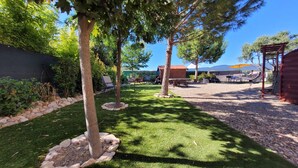 Un espacio exterior cuidado con césped, árboles, una pérgola de madera y una zona de estar con mesas y sillas.