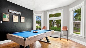Une table de billard dans une pièce dotée de grandes fenêtres, avec des tableaux encadrés accrochés au mur et un fauteuil marron.