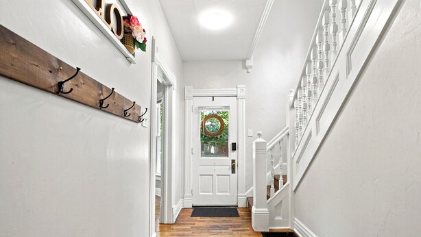 Un couloir avec un porte-manteau en bois, une porte blanche ornée d’une guirlande, et un escalier avec des rampes blanches.
