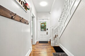 Un couloir avec un porte-manteau en bois, une porte blanche ornée d’une guirlande, et un escalier avec des rampes blanches.