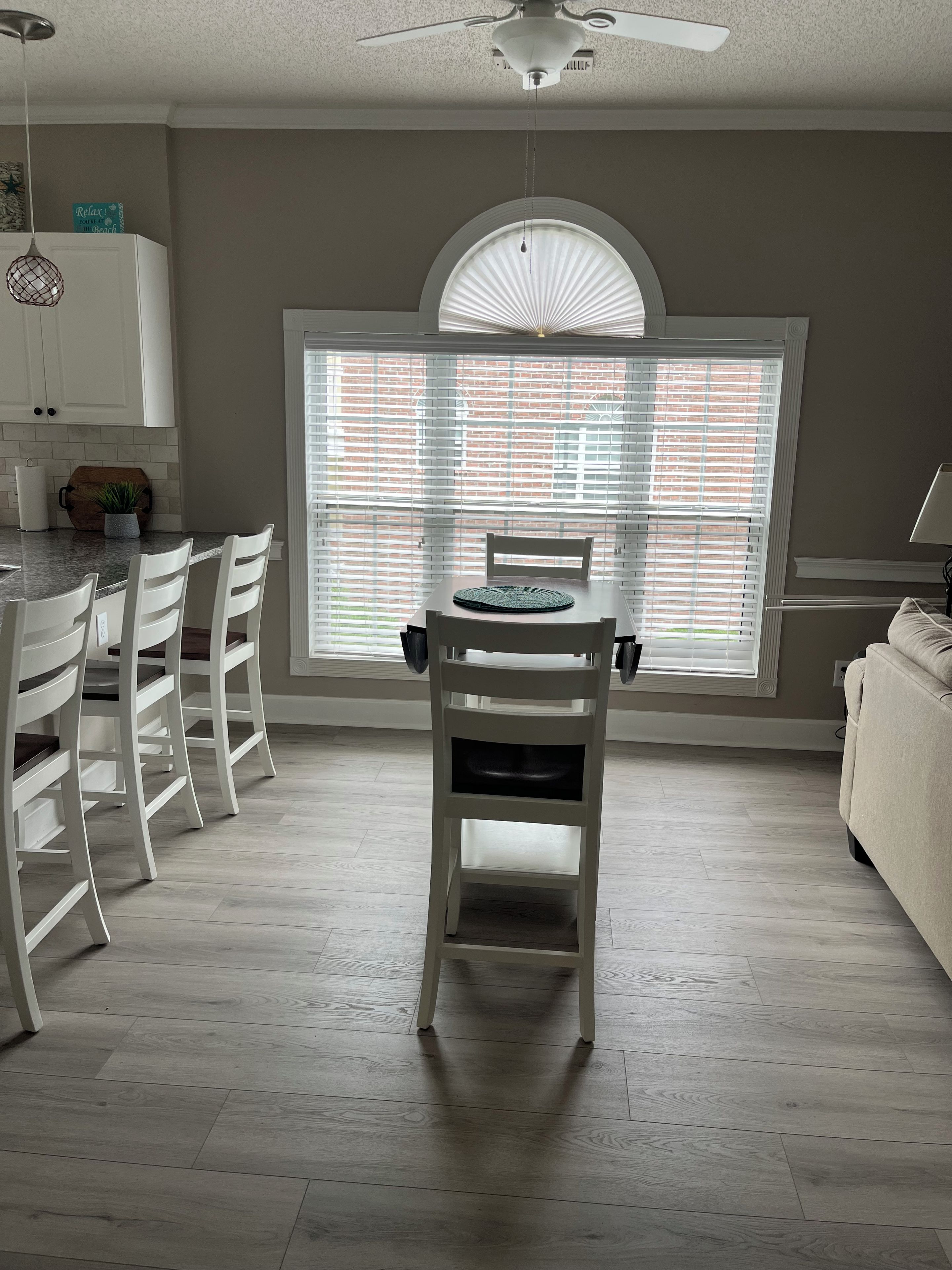 A dining area with a table and chairs, a window with blinds, a ceiling fan, and a kitchen area with cabinets and a plant.