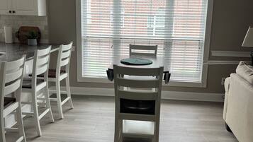 A dining area with a table and chairs, a window with blinds, a ceiling fan, and a kitchen area with cabinets and a plant.