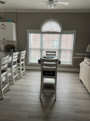 A dining area with a table and chairs, a window with blinds, a ceiling fan, and a kitchen area with cabinets and a plant. - Charming 2-bedroom, 2 bath condo with AC, WiFi in welcoming Little River. (Little River)