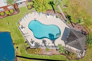An aerial view of a swimming pool with a surrounding deck, palm trees, and a building in the background.