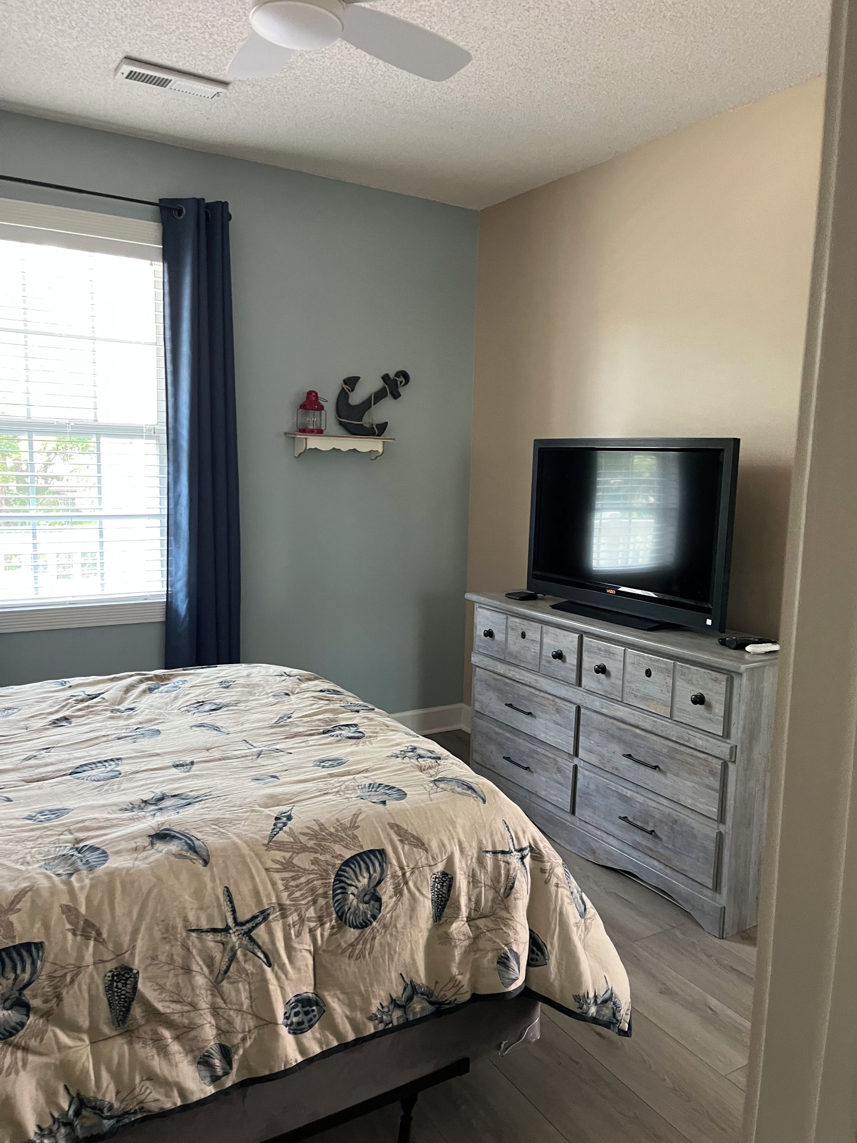 A bedroom with a bed, a dresser, a television, a window with curtains, and a wall-mounted decoration.