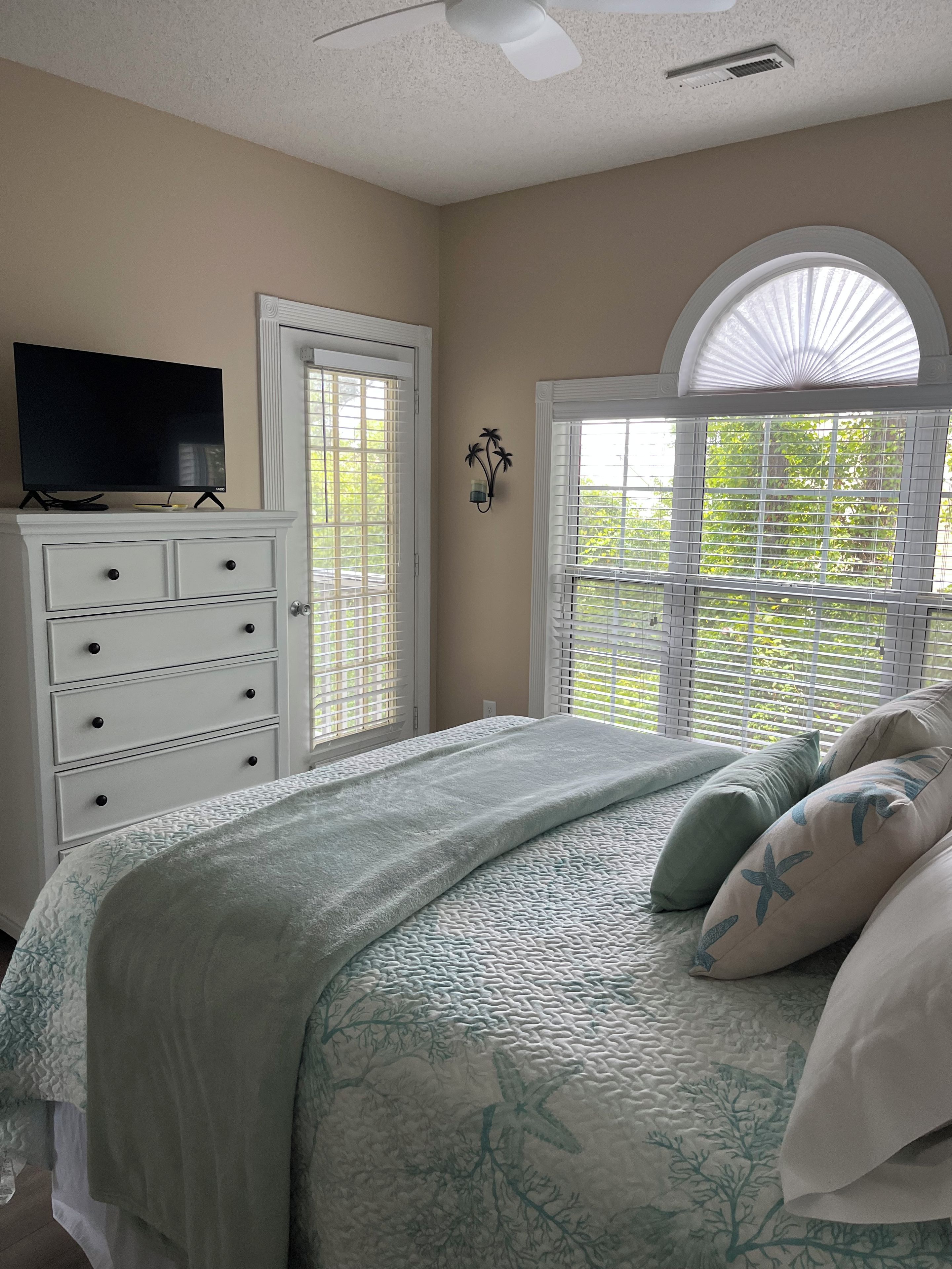 A bedroom with a bed, a dresser, a television on top, and a window with blinds.