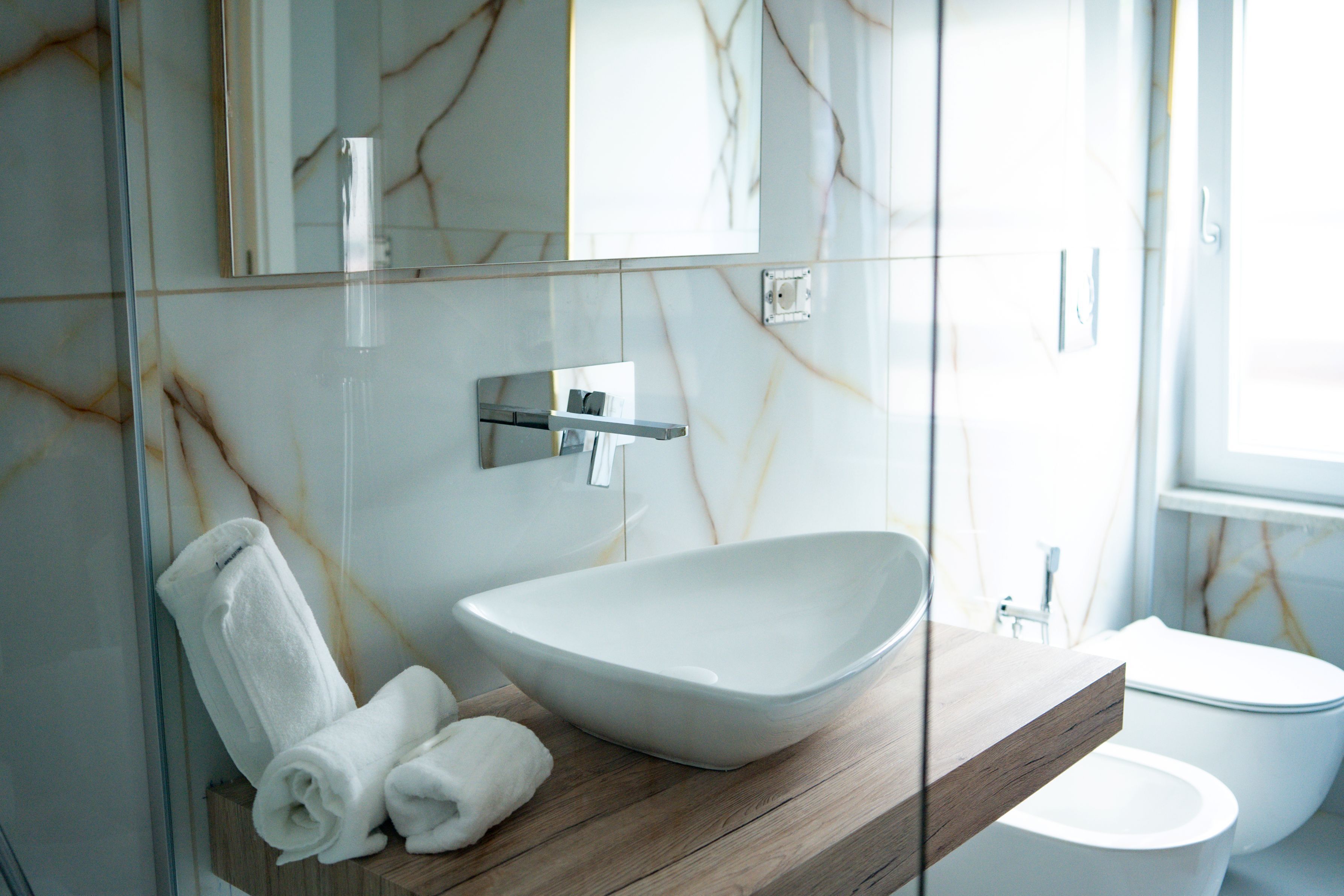 Un baño con pared de mármol, tocador de madera, lavamanos blanco y un inodoro.