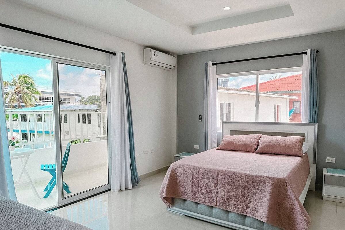 Un dormitorio moderno con una cama grande, un balcón con vistas a edificios y un equipo de aire acondicionado.