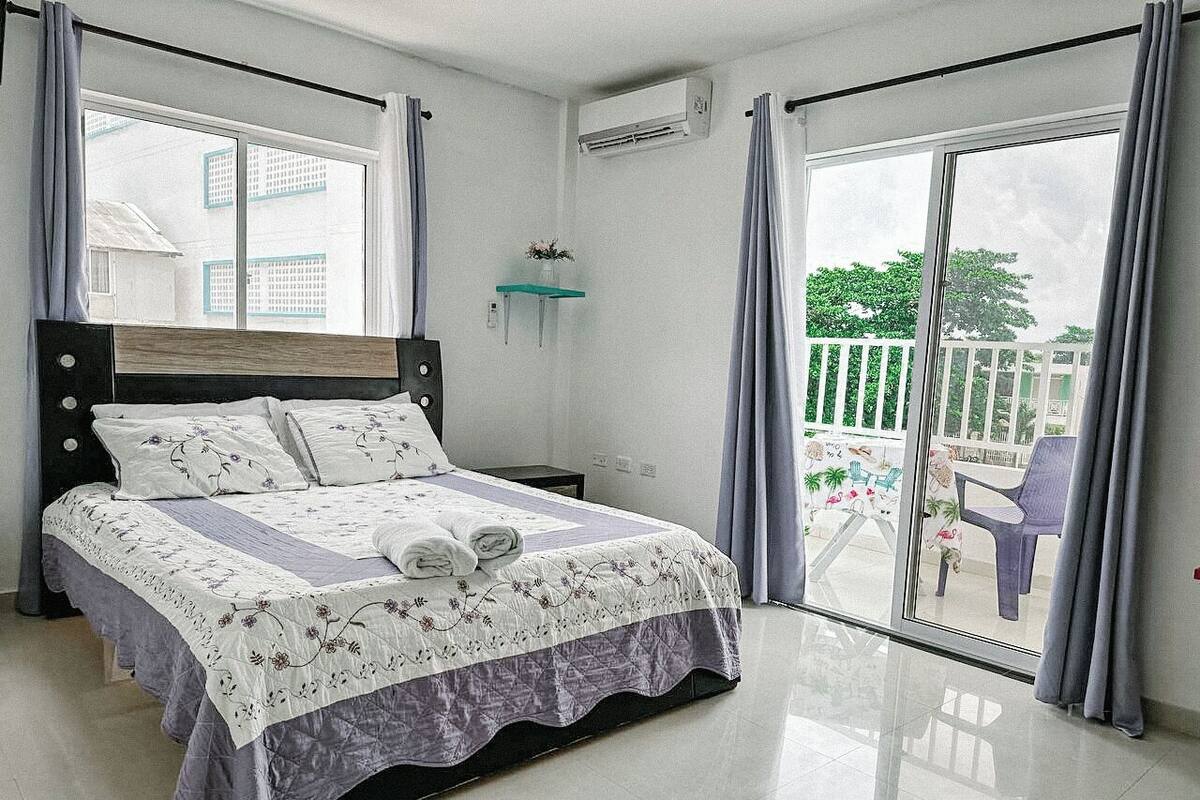 Un dormitorio con cama, mesita de noche, ventana y balcón con sillas.