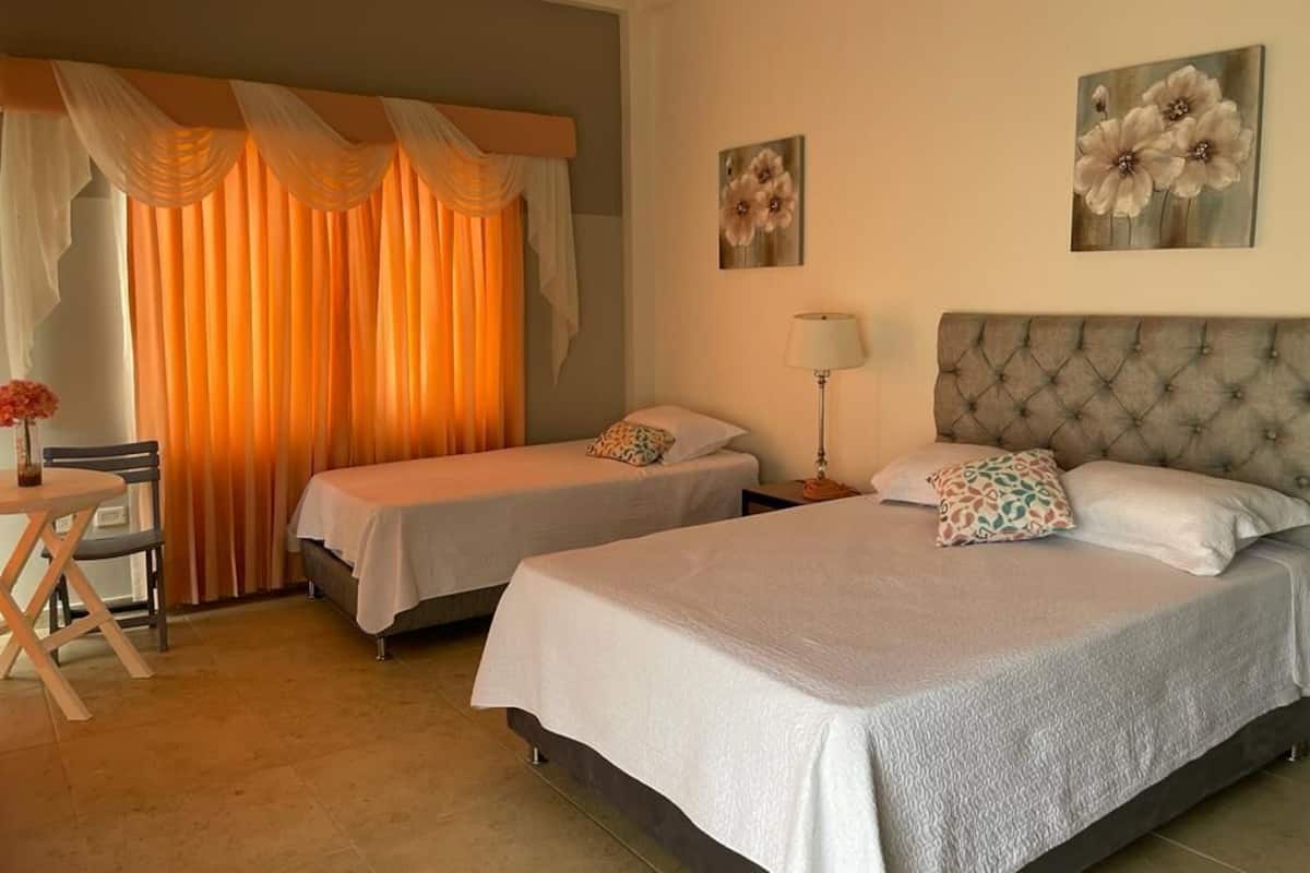 Habitación de hotel con dos camas individuales, un comedor con mesa y sillas, y un ventanal con cortinas naranjas.